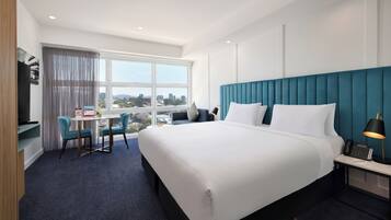 Quarto de hotel com uma cama grande, uma escrivaninha com cadeira, televisão e janela com vista para a cidade.