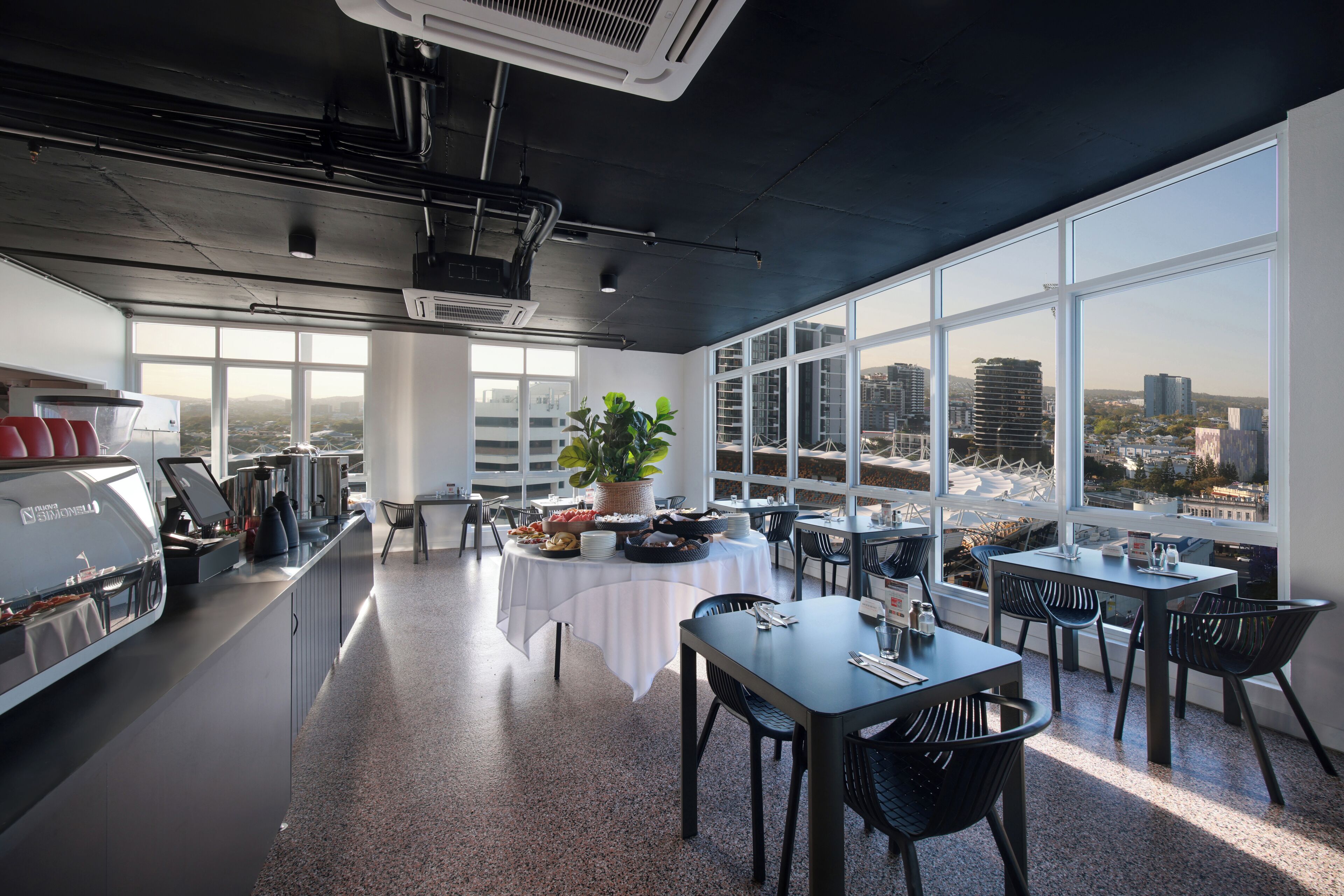 A modern dining area with a view of the city, featuring a buffet setup, seating arrangements, and a large window.