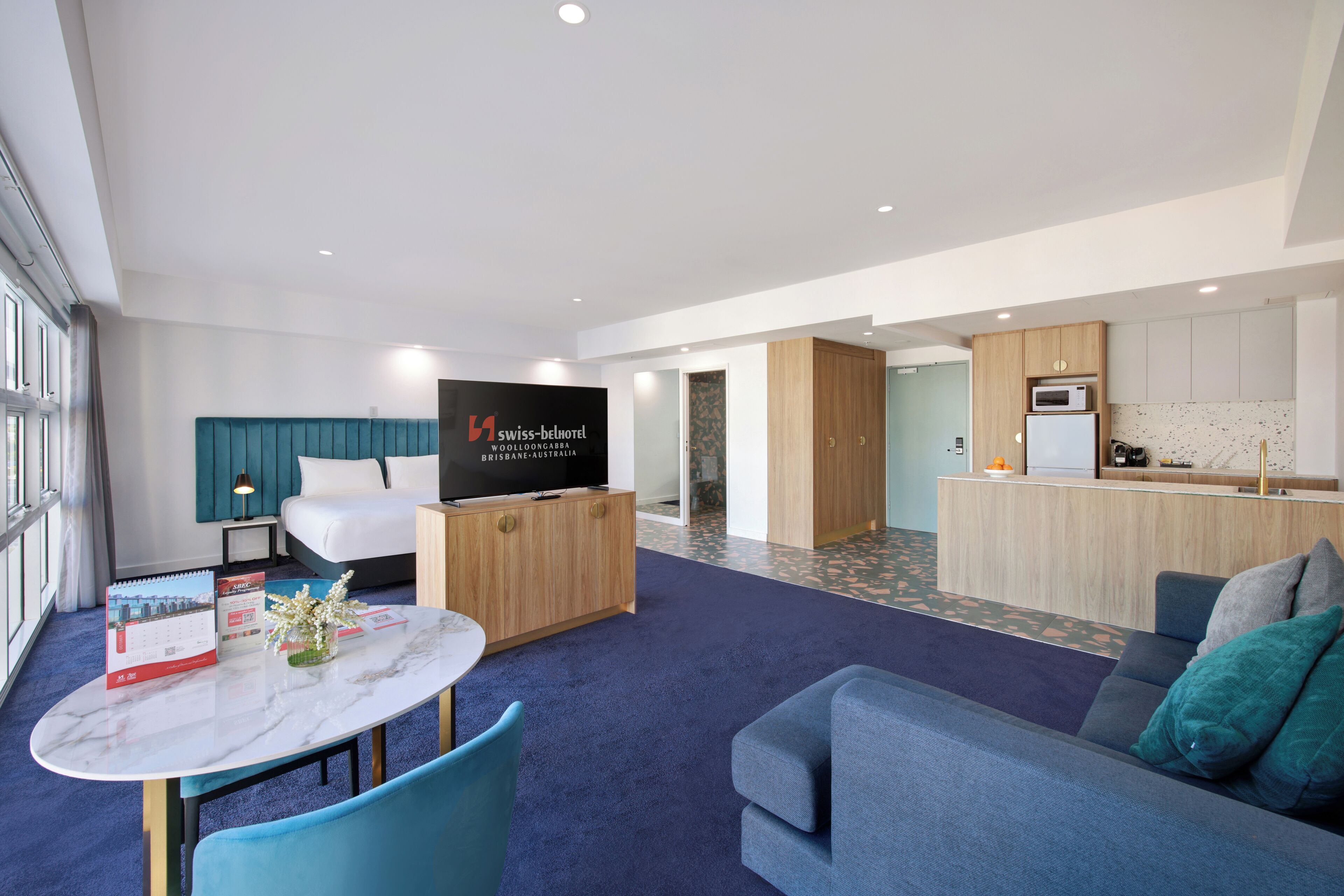 A modern hotel room with a large bed, a TV, a dining area with a table and chairs, a kitchen area, and a living area with a sofa.
