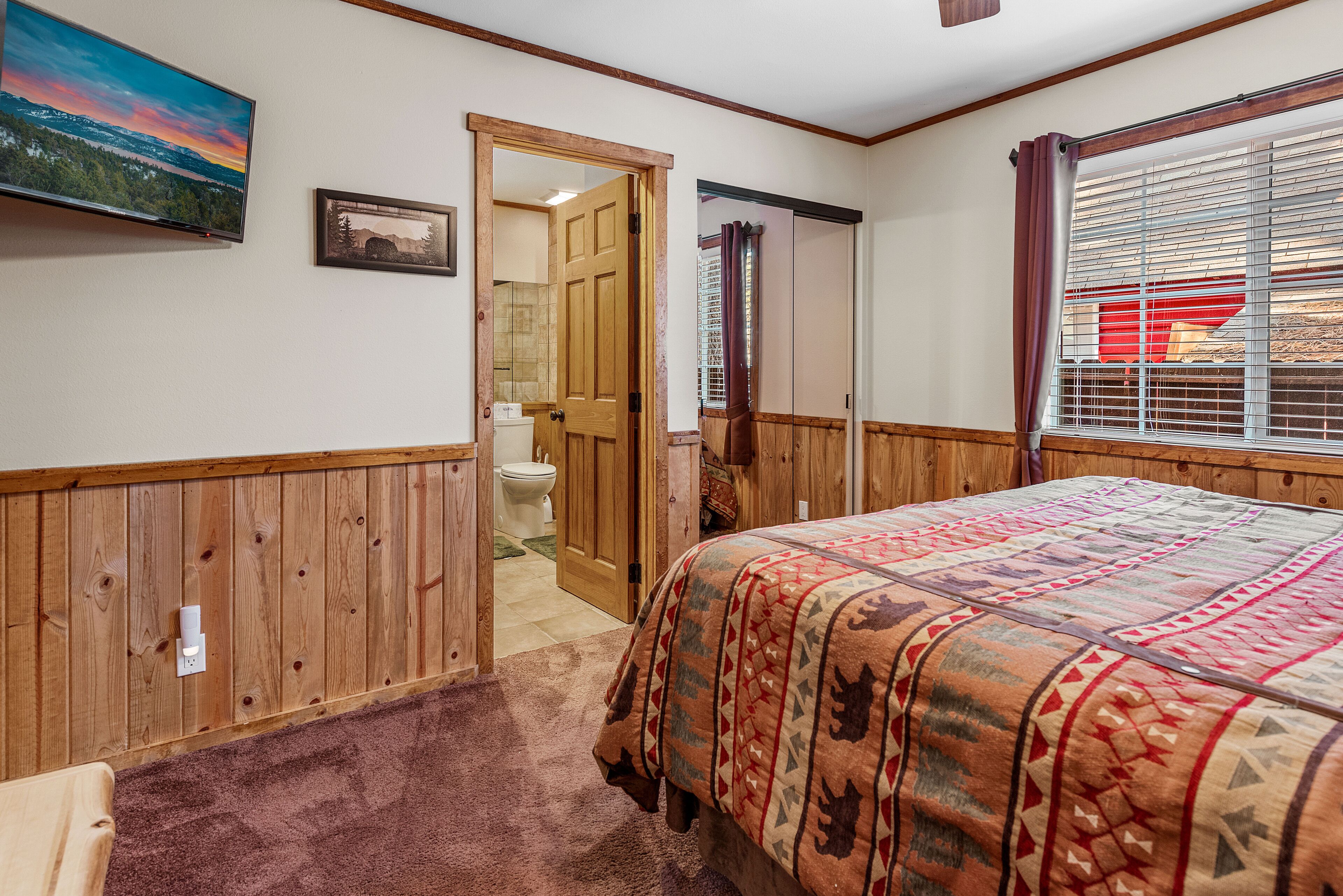 Un dormitorio con pared de madera empapelada, una cama con manta estampada, un televisor montado en la pared y una ventana con persianas.