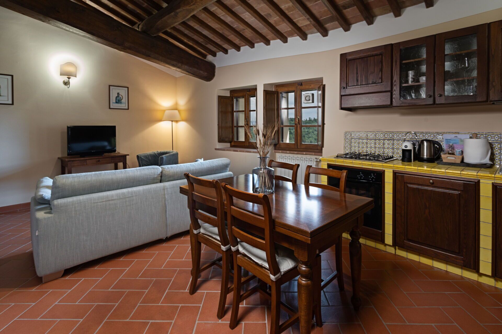 A cozy living area with a sofa, TV, and dining table with chairs. A kitchen with cabinets and a stove is visible.