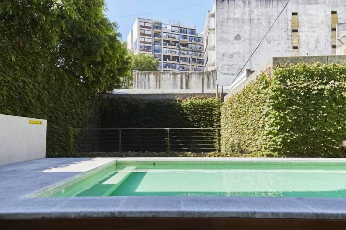 A swimming pool with a concrete deck, surrounded by greenery and a building in the background.