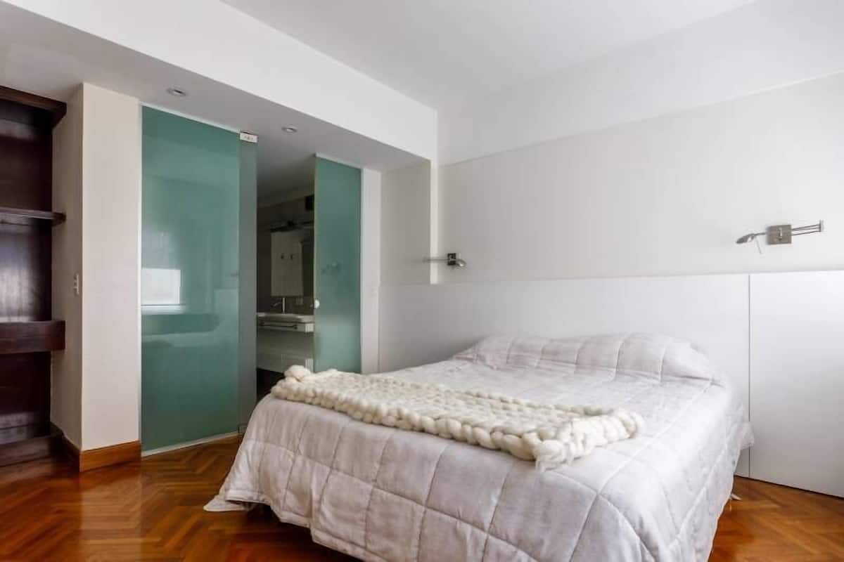 A bedroom with a bed, a wooden wardrobe, and a glass door leading to a bathroom.