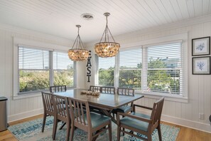 Uma área de refeições com mesa e cadeiras de madeira, janelas amplas com persianas e duas luminárias pendentes.