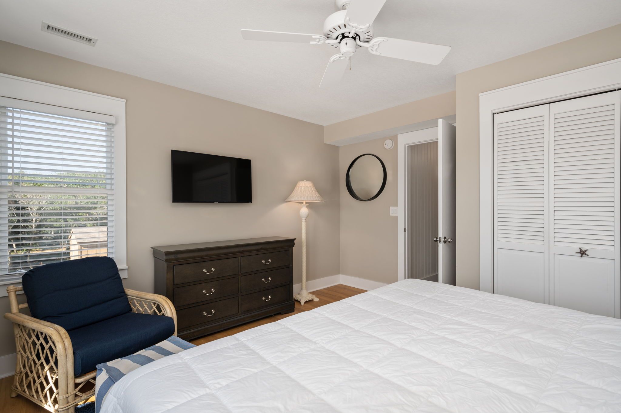 A bedroom with a bed, dresser, chair, ceiling fan, TV, lamp, and a door.