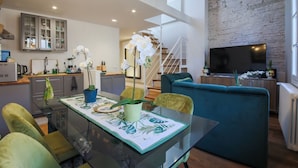 Un moderno salón con mesa de vidrio, sillas verdes, sofá azul, cocina con armarios, una escalera y un televisor de pantalla plana.