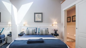 Un dormitorio con una cama grande, dos mesitas de noche, una mesita auxiliar, un cuadro enmarcado en la pared y una puerta que da a otra habitación.