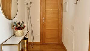 Un passadís amb una porta de fusta, un mirall, una taula petita amb un cistell de flors i una escala metàl·lica.