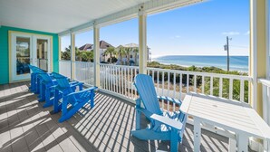 A covered outdoor area with blue Adirondack chairs and a table, overlooking a beach with houses and the ocean. - Surfer Dog 4 Bedroom Home by RedAwning (Panama City Beach)