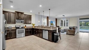 A modern kitchen with dark wood cabinets, stainless steel appliances, and a large island. There is a dining area with a sofa and a view of the outdoors. - Championsgate Resort 9 Bedroom Private Pool Home (Davenport)