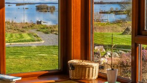 Blick durch ein Fenster auf einen See mit einer Wiese und einem Korbkörbchen im Fensterbrett.