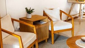 A room with two wooden chairs with white cushions, a small wooden side table with a plant, and a round wooden table with a bowl.