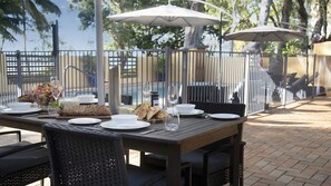 An outdoor dining area with a table set for two, surrounded by a metal fence, and umbrellas providing shade.