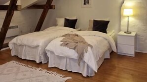 Et soveværelse med skråvæg, træbjælker, en seng med hvide sengetæger, et sengebord med lampe og et tæppe på gulvet.