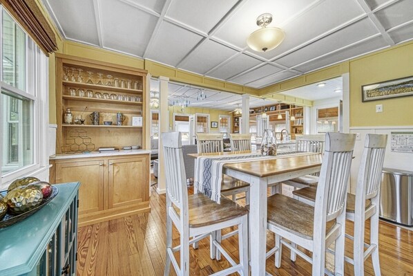 A spacious kitchen with wooden cabinets, a dining table, and chairs. There are shelves with glassware and a window providing natural light.