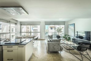 A modern kitchen with a central island, a dining area with a table and chairs, a living room with a sofa and a TV, and large windows offering a view of the city.