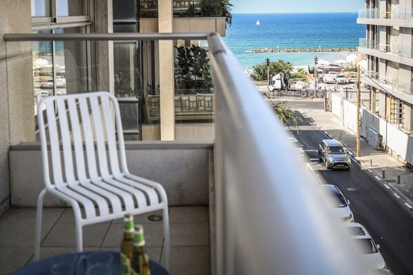 A balcony with a white chair, a bottle, and a view of the sea and a road with parked cars. - Mamad Luminous 3BR Escape with Balcony by SeaNRent (Tel Aviv)