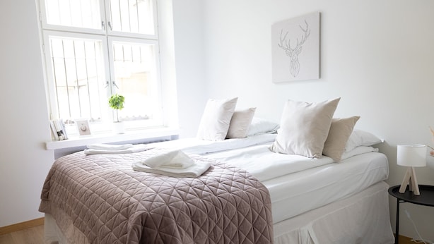 Et soveværelse med seng, puder, et natbord, en lampe og et vindue med persienner.