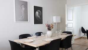 En spisestue med et træbord dækket til seks personer, sorte stole, en vase med tørrede blomster og to billeder i rammer på væggen.