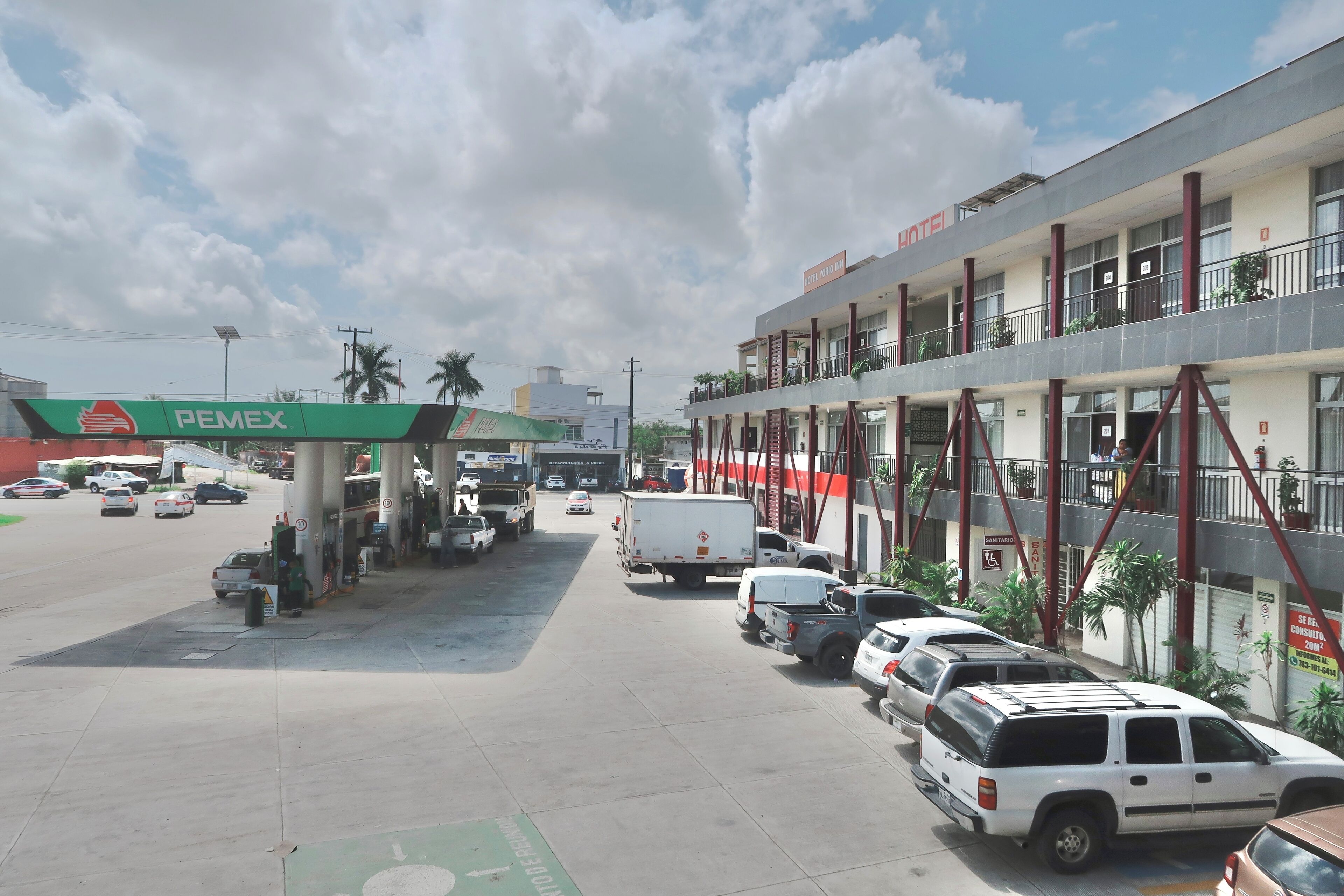 Un hotel con balcón en el piso superior, una estación de servicio con la marca PEMEX y varios vehículos estacionados.