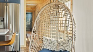 Eine Holzbalkendecke mit eingelassener Beleuchtung, ein hängender Korbstuhl mit blauen Kissen, ein Edelstahlkühlschrank und ein Holzboden.