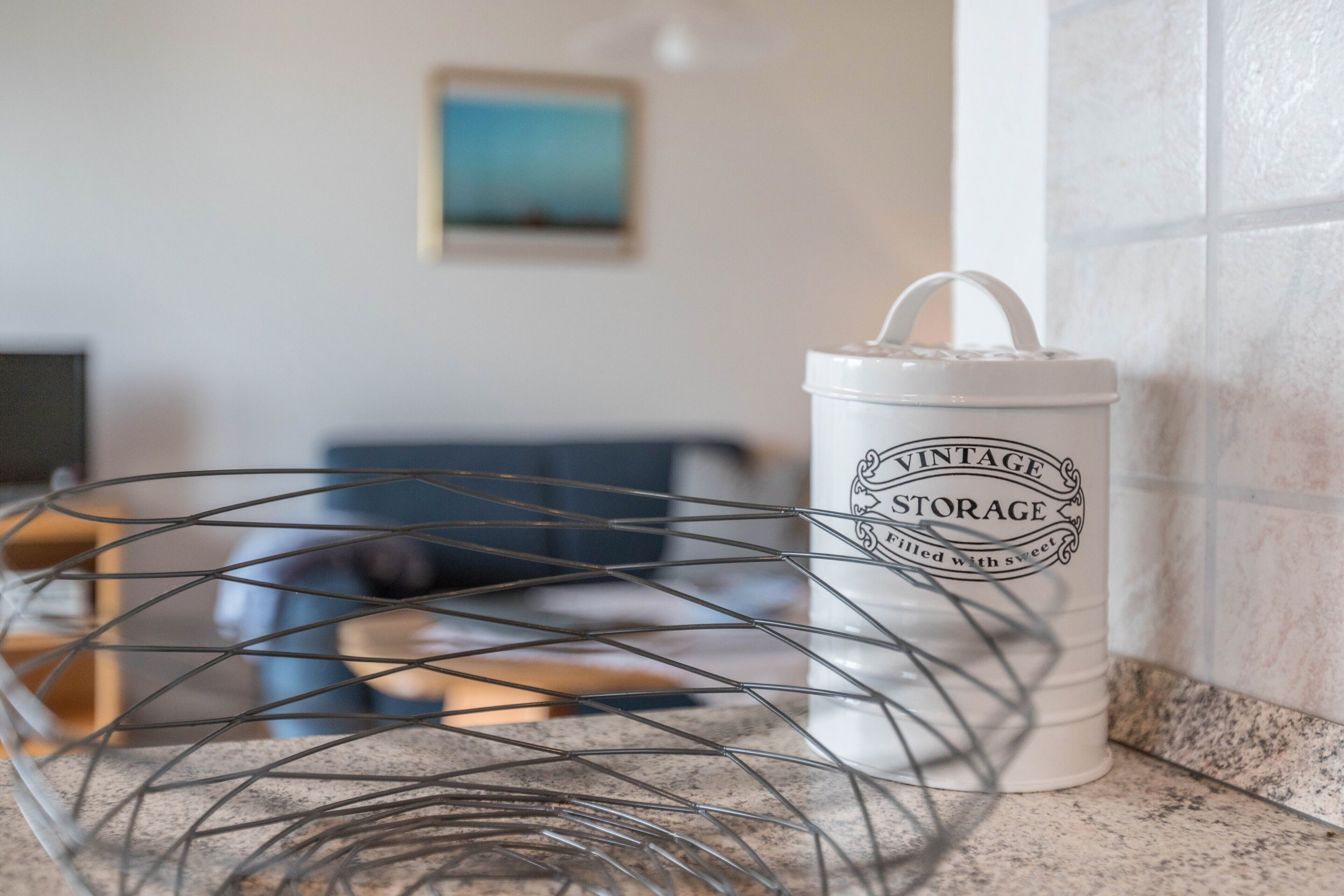 A kitchen counter with a white storage container labeled 'VINTAGE STORAGE' and a metal wire basket.