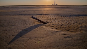 Una playa arenosa con un poste de madera y una estructura distante, posiblemente un faro, bajo un cielo despejado.
