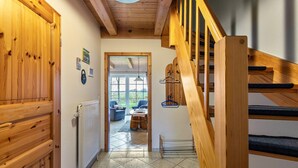 Escada de madeira que dá para um segundo andar, corredor com radiador e vista para uma sala de estar com mesa e cadeiras.