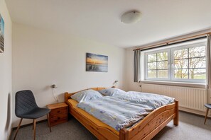 A bedroom with a wooden bed, a chair, a nightstand, a window with curtains, and a wall-mounted picture.