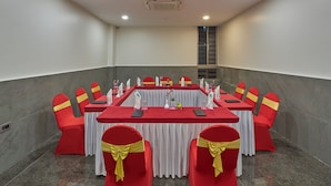 Uma sala de conferências com uma mesa comprida, cadeiras vermelhas com fitas douradas, toalhas de mesa brancas e uma cortina branca na frente.