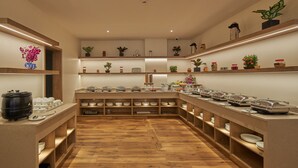 Disposição de buffet com várias estações de serviço, piso de madeira e prateleiras com plantas decorativas e potes.