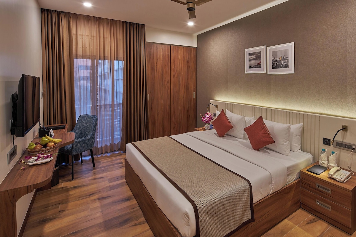 A hotel room with a bed, bedside table, desk with a chair, a television, and a window with curtains.