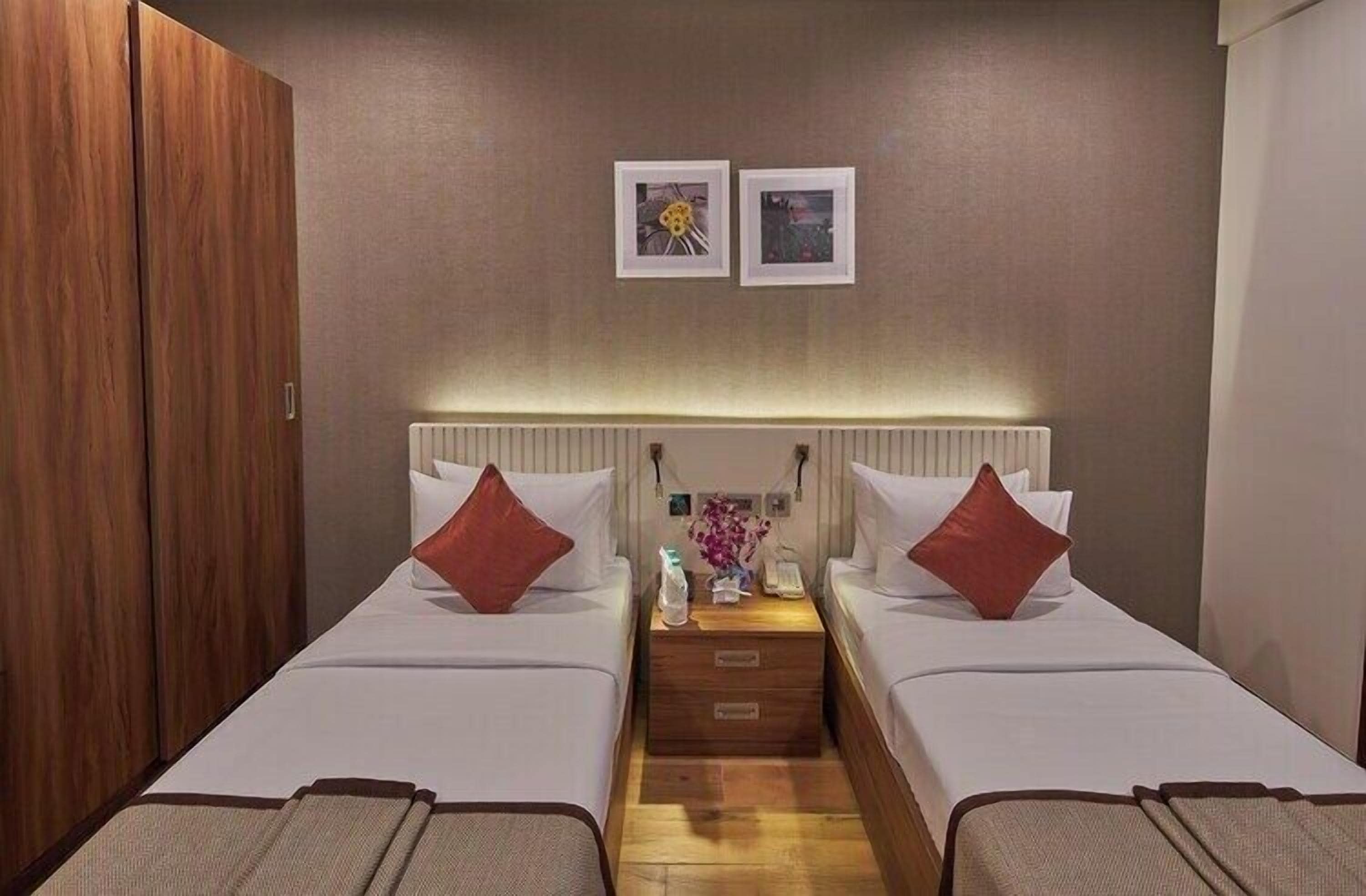 Habitación de hotel con dos camas, un armario de madera, una mesita de noche con lámpara y dos cuadros en la pared.