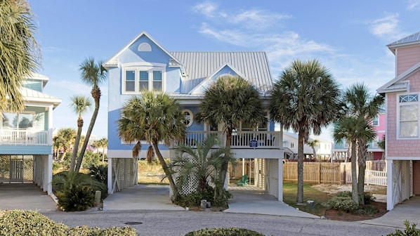 A two-story house with a balcony, surrounded by palm trees and a wooden fence.