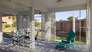 A covered patio area with a table and chairs, a green Adirondack chair, and a wooden fence in the background.