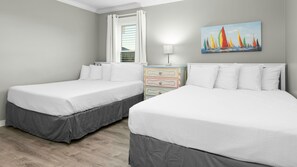 A hotel room with two beds, a window with white curtains, a colorful dresser, and a painting of sailboats on the wall.