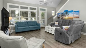 Een woonkamer met een blauwe bank, een grijze bank, een witte fauteuil en een witte salontafel. Links is een televisie en erboven een plafondventilator.