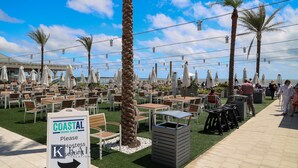 Een terras aan zee met tafels en stoelen, palmbomen en een bord dat een kioskzone aangeeft.