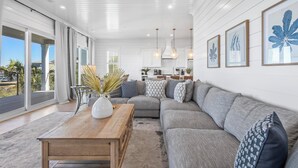 Uma sala de estar moderna com um sofá seccional cinza, uma mesa de centro de madeira e um ventilador de teto. Há gravuras botânicas emolduradas na parede e uma vista do exterior através de grandes portas de vidro deslizantes.