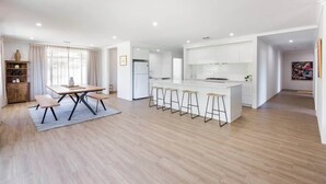 A modern kitchen with white cabinets, a central island with bar stools, and a dining area with a table and benches. - Wildgreen Villa—Lakeside Hideaway in Winery Estate (Yallingup Siding)