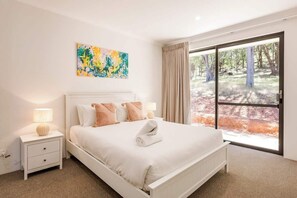 A bedroom with a large bed, a nightstand with a lamp, a painting on the wall, and a view of a forest through a sliding door.