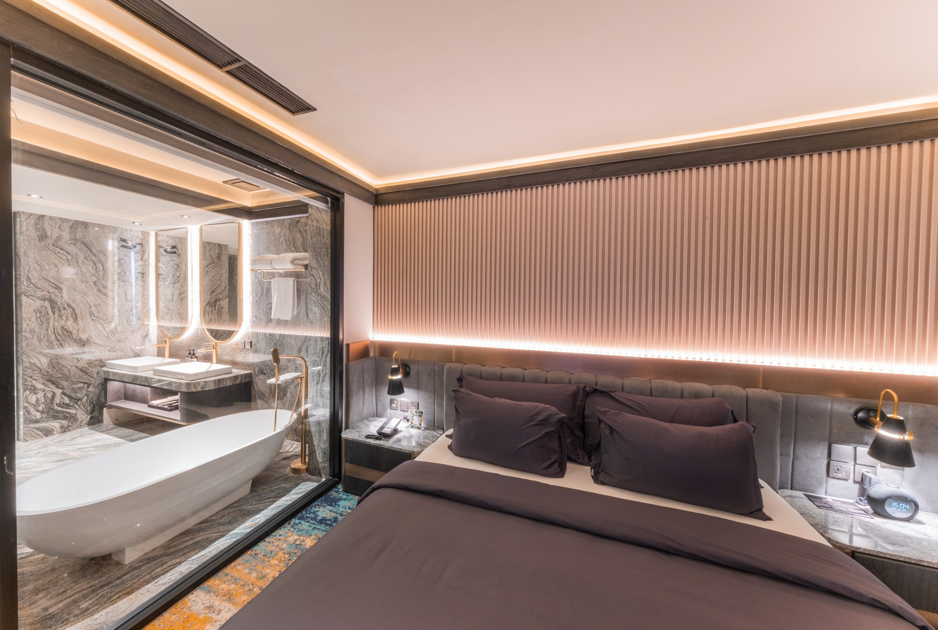 A modern hotel room with a large bathtub, a bed with dark bedding, and a bedside table with a lamp and alarm clock.