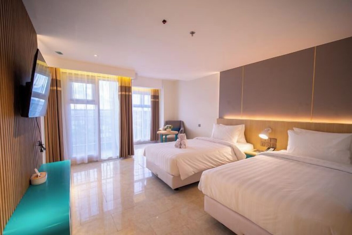 A hotel room with two beds, a TV mounted on the wall, a small seating area, and a window with curtains.
