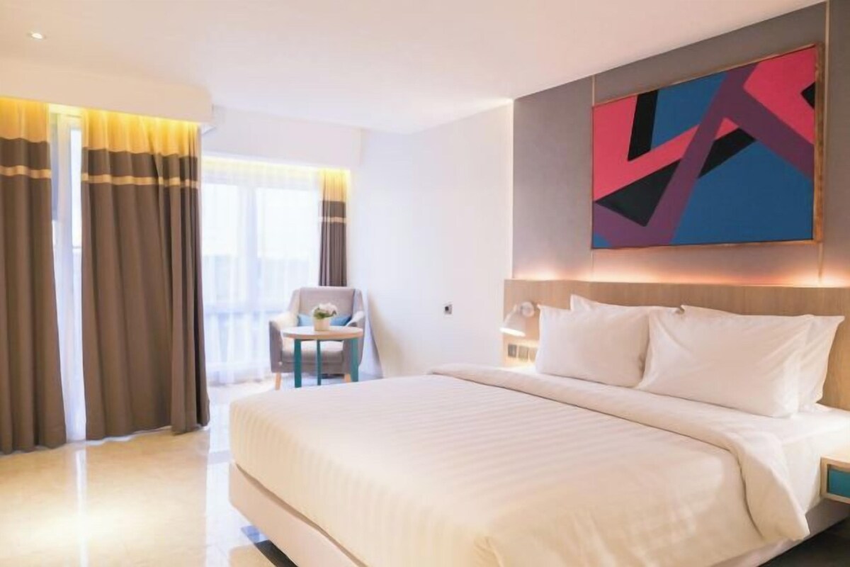 A hotel room with a large bed, a chair, a small table, and a painting on the wall.