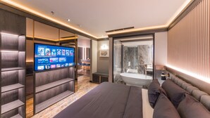 Een moderne hotelkamer met een groot bed, een televisie waarop een menu wordt weergegeven, een badkamer met een douchecabine van glas en een zithoek met een bank en een tafel.