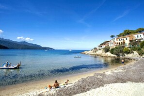 A coastal beach with clear waters, boats, and buildings along the shore.