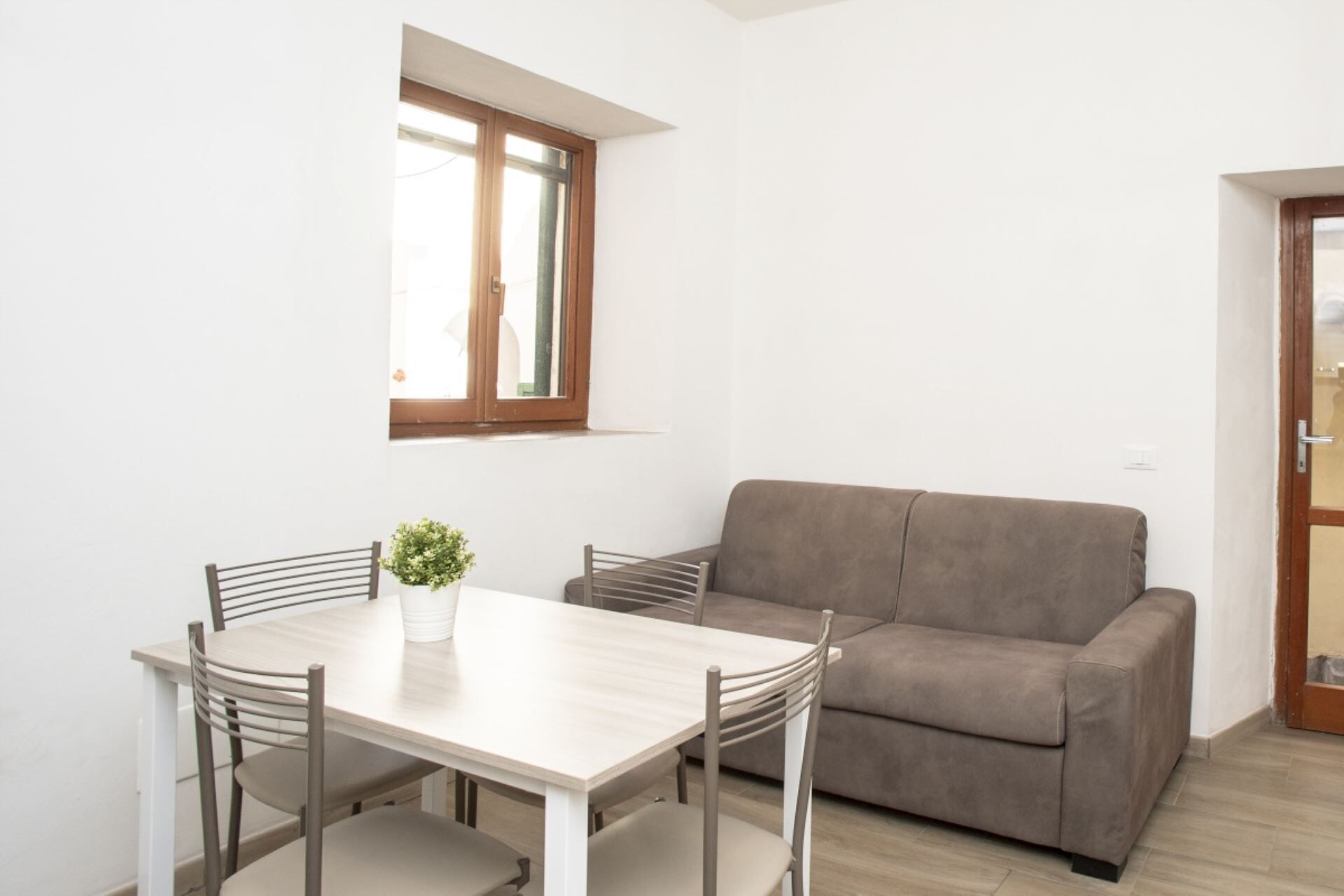 A small, bright room with a wooden-framed window, a beige sofa, and a wooden dining table with four chairs.