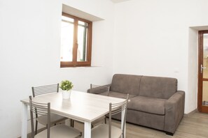 A small, bright room with a wooden-framed window, a beige sofa, and a wooden dining table with four chairs.