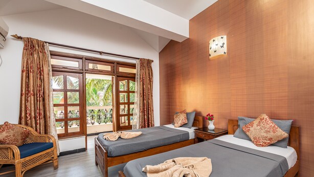 A hotel room with two beds, a wooden wall, a window with curtains, a wicker chair, and a nightstand with a vase. - Villa Calangute Phase 1 (Calangute)