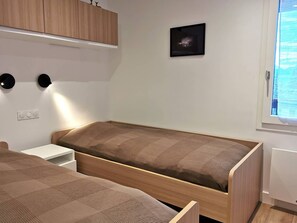 Ein kleiner, moderner Raum mit zwei Einzelbetten, einem Holzschrank, einem Wandbild und einem Fenster mit Jalousien.